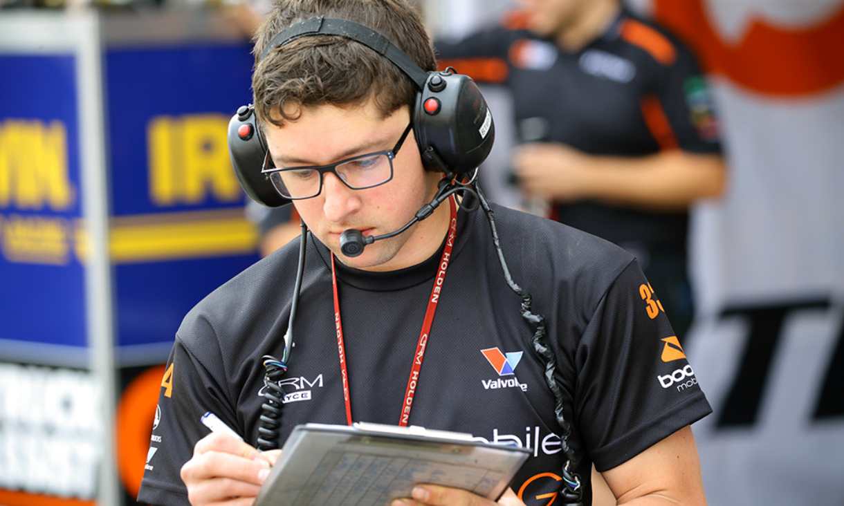 Close up of Stefan. He has short brown hair and glasses. He is wearing a navy team shirt and a headset. He has a clipboard and pen in his hand.