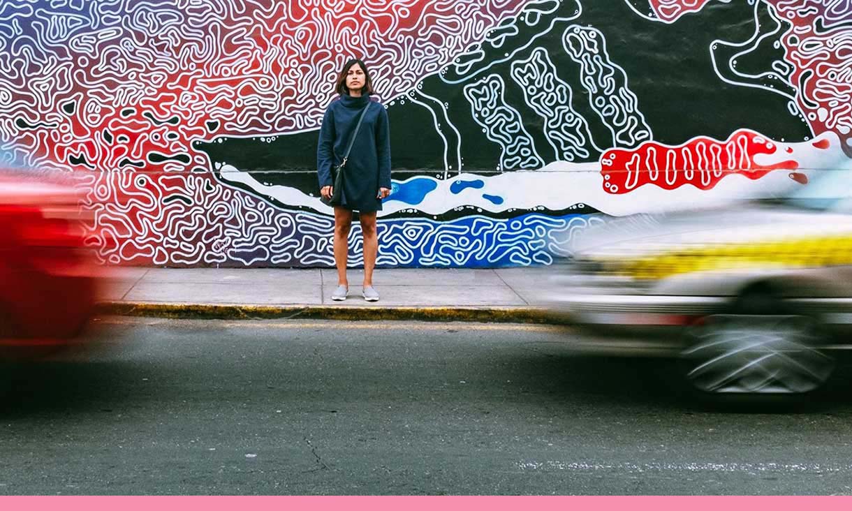 A person stands against a wall of graffiti on a footpath while cars drive past.