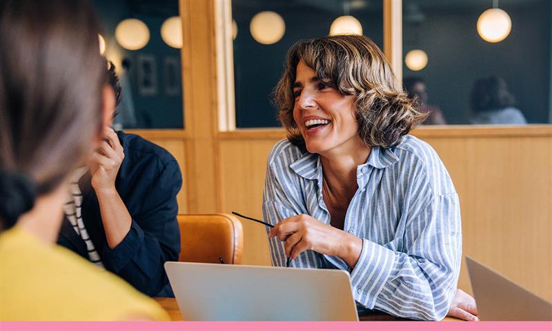 A group of people in a meeting, one female smiling and looking towards the other two females with a laptop at a shared desk.