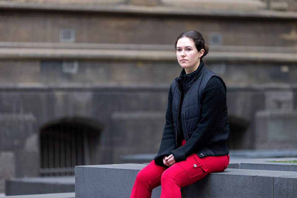 A girl sitting outside with a concrete building behind her. She wears a black jumper and red pants.