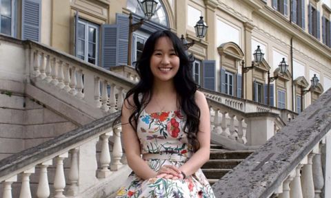 ugenie is a young Asian woman. She has long dark hair and is wearing a floral sundress. She is sitting on the banister of an old stone staircase at the front of a building. 