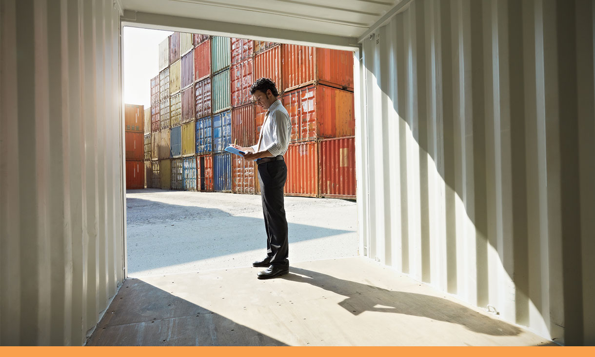 A man stands next to shipping containers and makes notes on paper.