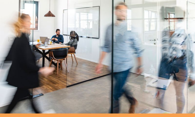 Two people in a meeting room while others walk past.
