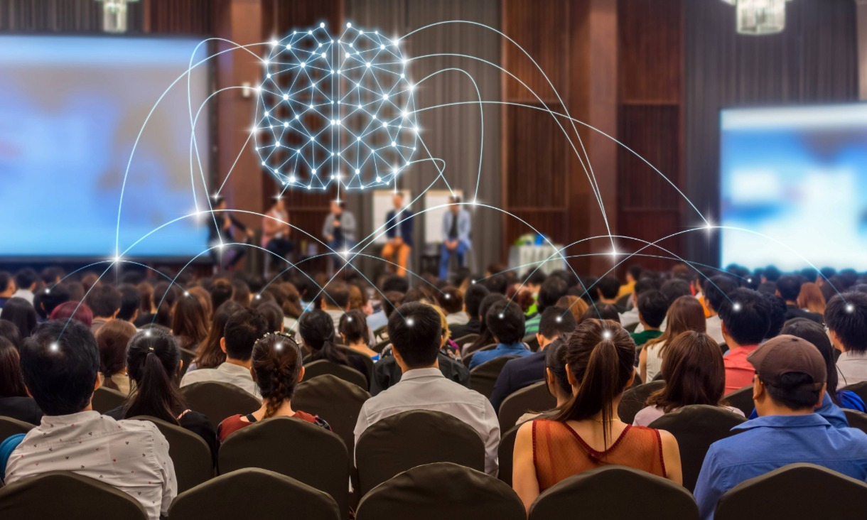 Large audience facing a panel of speakers at a conference, with a digital brain graphic and network lines symbolising connectivity and technology.