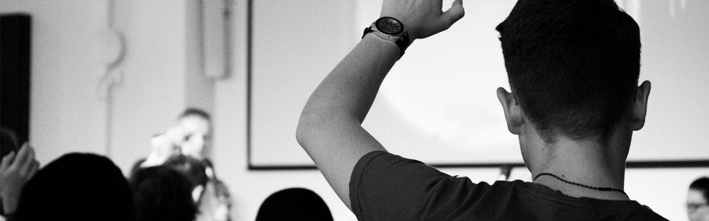 Black and white image from behind a student raising their hand in a lecture
