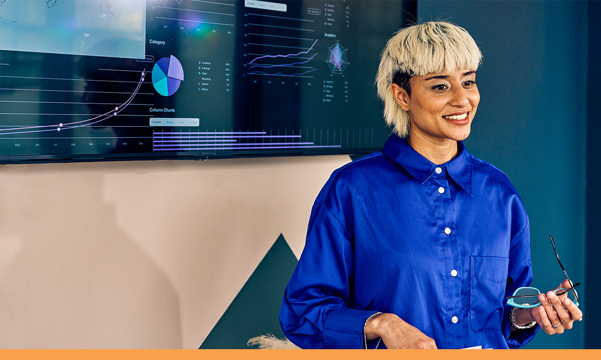 A woman stands in front of screen with data and tables displayed.