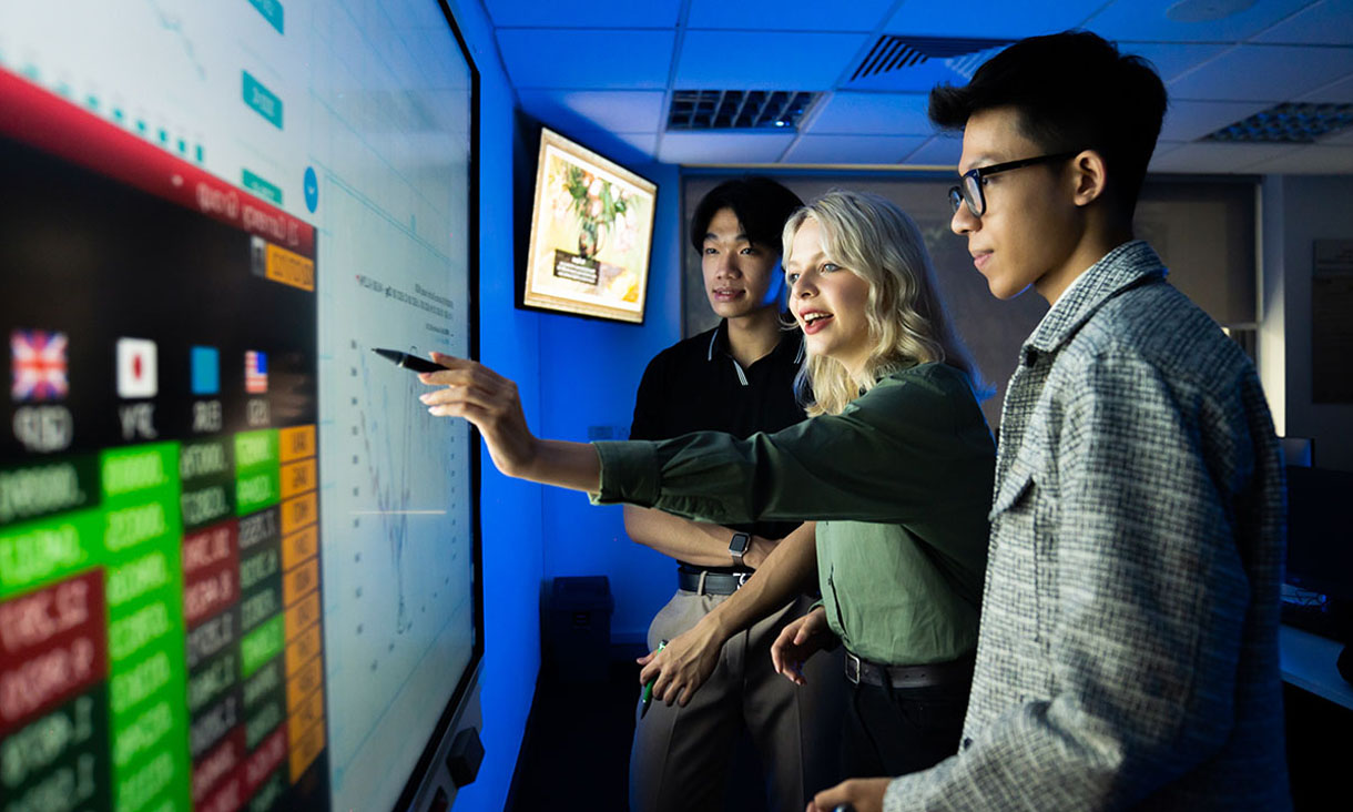 A woman points at a graph on a large screen while two men listen to her.