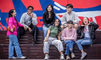 A group of seven students are sitting on a outdoor stairs on a university campus, looking at the camera and smiling.