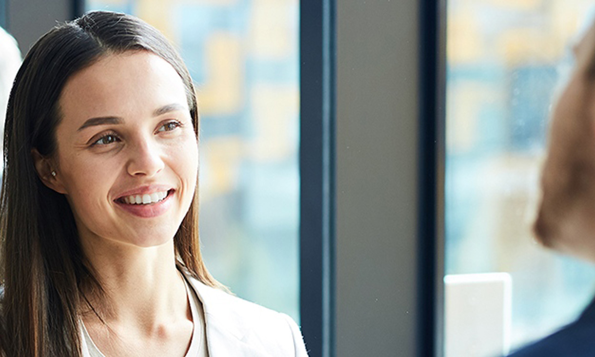 A woman in focus is smiling while talking to a man in an office.