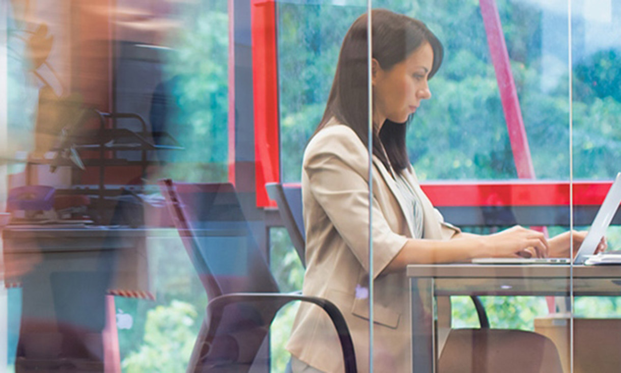 A woman works in an office while people walk past outside.